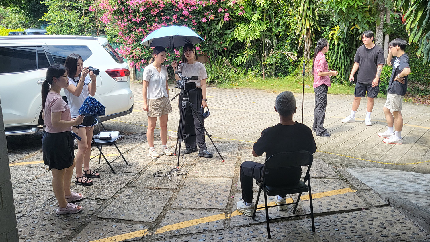 Seven students with various filming equipment stand in small groups around a seated Dr. Urbi for an interview.